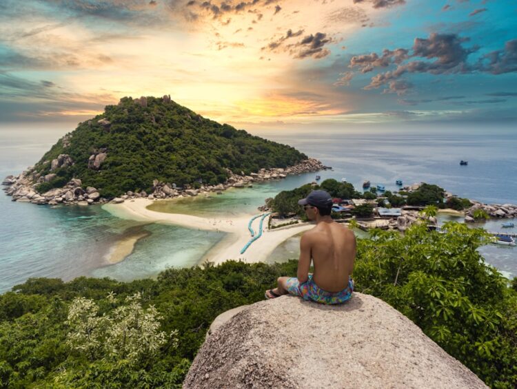 topless man sitting on cliff rock viewing Ko Nang Yuan isalnd, houses, and buildings under white and yellow sky